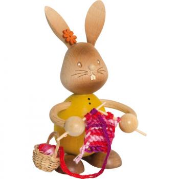 Wooden rabbit Stupsi with a yellow shirt holds a brush in his hand and paints the red Easter egg in front of him.