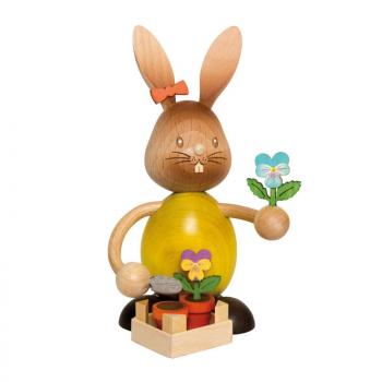 Wooden rabbit Stupsi with a yellow shirt and a pansy in his hand. In front of him is a pot planted with a pansy.