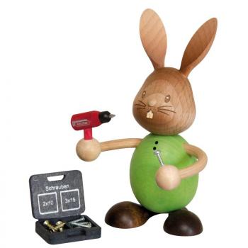 Stupsi the wooden rabbit in a green shirt is holding a cordless screwdriver and a screw in his hand. The toolbox is lying on the floor in front of him.