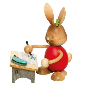 Wooden rabbit Stupsi with a red shirt is standing at his school desk, solving a problem and writing as he goes. He is holding a pencil in his hand.