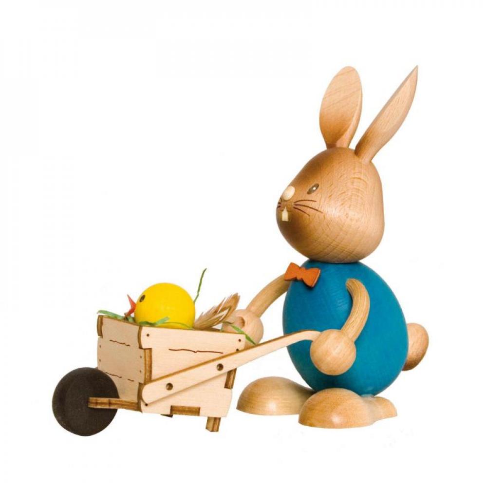 Wooden rabbit Stupsi with a blue shirt is pushing a wheelbarrow in front of him. A little chick is sitting in the cart and looking out.