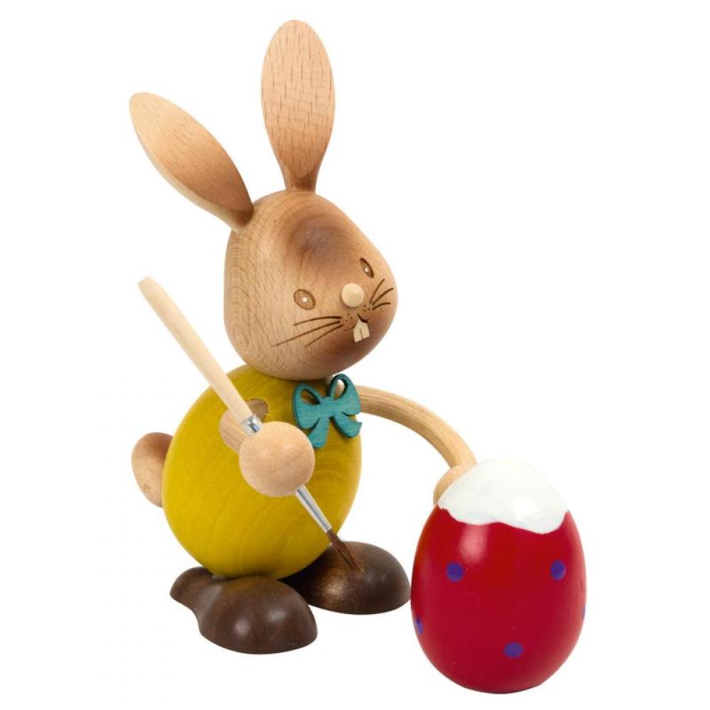 Holzhase Stupsi mit gelben Shirt hält einen Pinsel in der Hand und bemalt das rote Osterei vor sich.