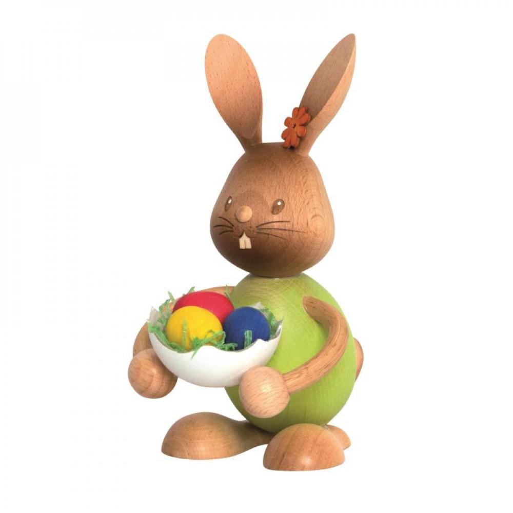 Wooden rabbit Stupsi with a green shirt holds a large eggshell in his hand with three colored eggs in it.