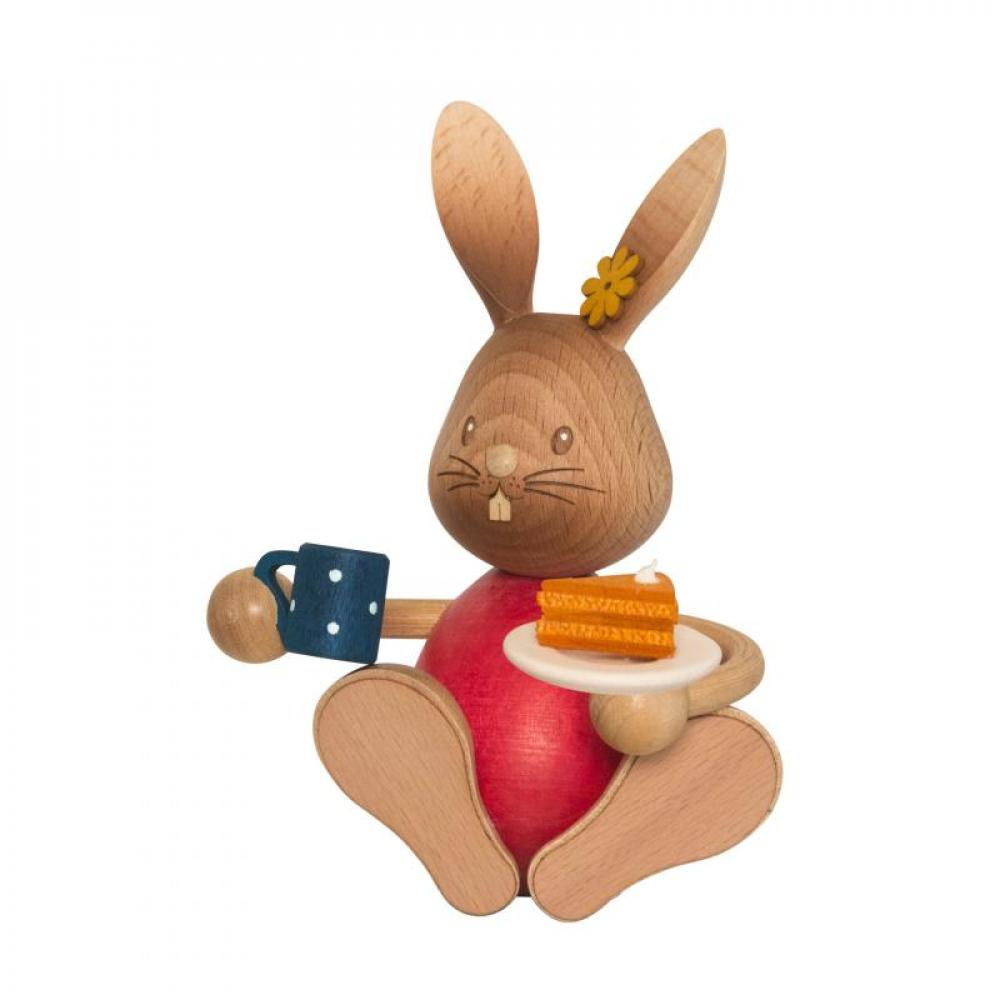 Wooden rabbit Stupsi with red shirt holds a cup in one hand and a piece of cake on a plate in the other.