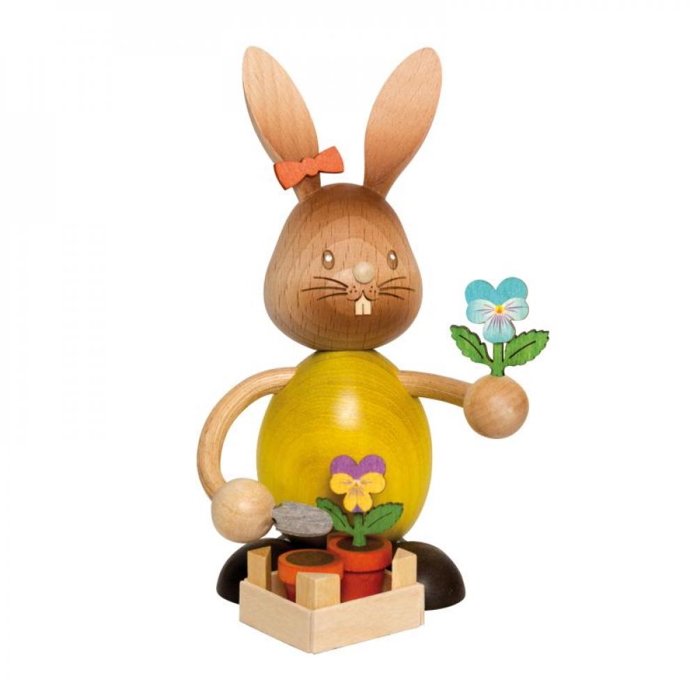 Wooden rabbit Stupsi with a yellow shirt and a pansy in his hand. In front of him is a pot planted with a pansy.