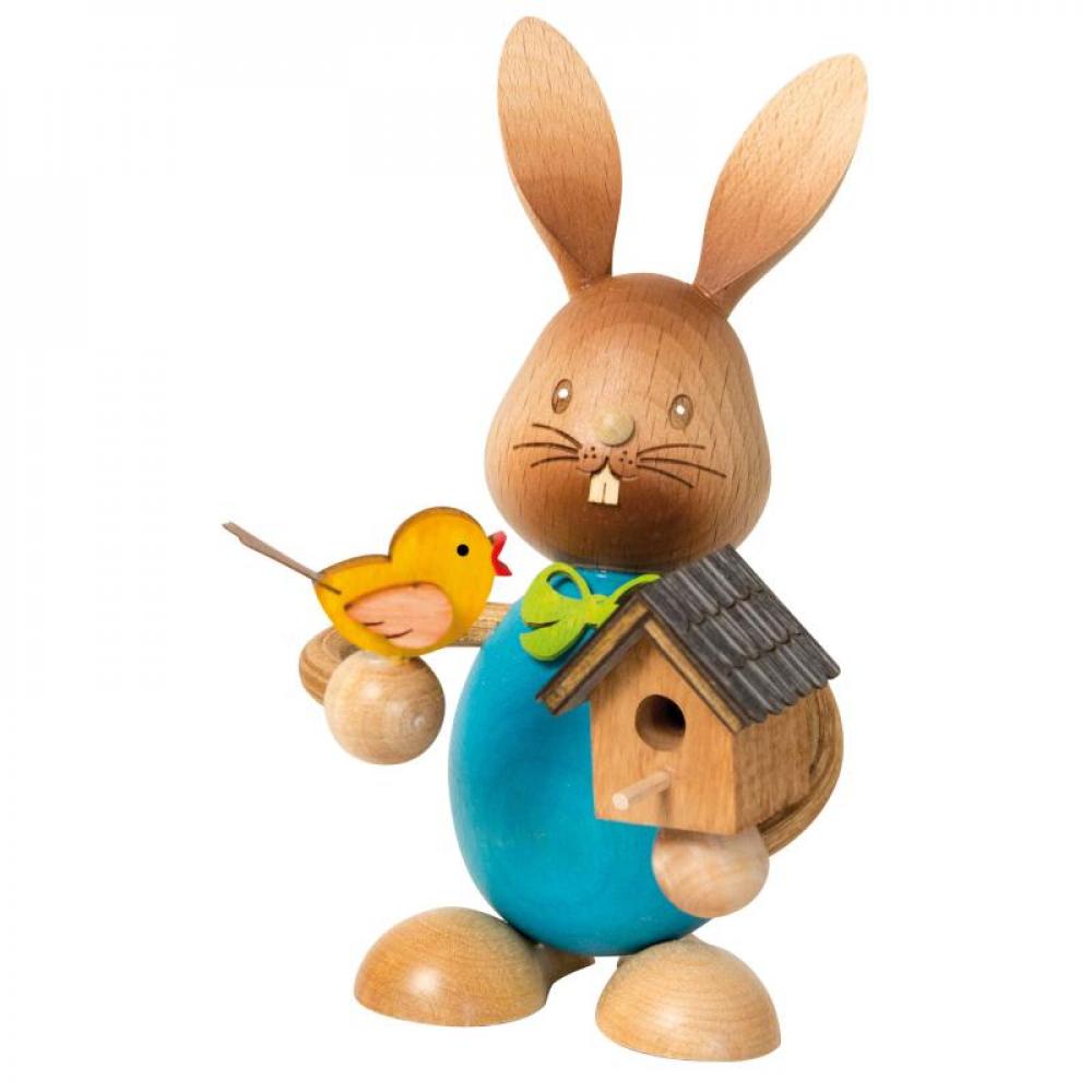 Wooden rabbit Stupsi with a blue shirt and holding a birdhouse in one hand and a yellow bird sitting on his other hand.