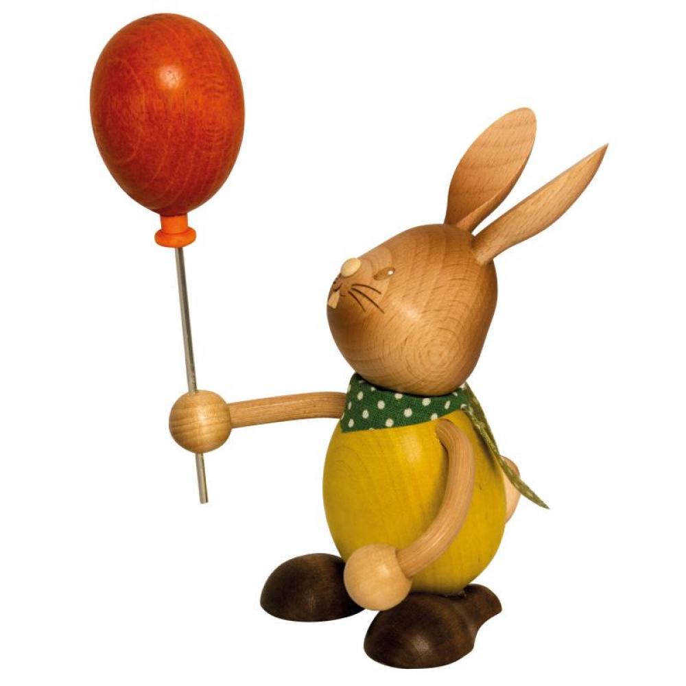 Wooden rabbit Stupsi with a yellow shirt holds a brush in his hand and paints the red Easter egg in front of him.