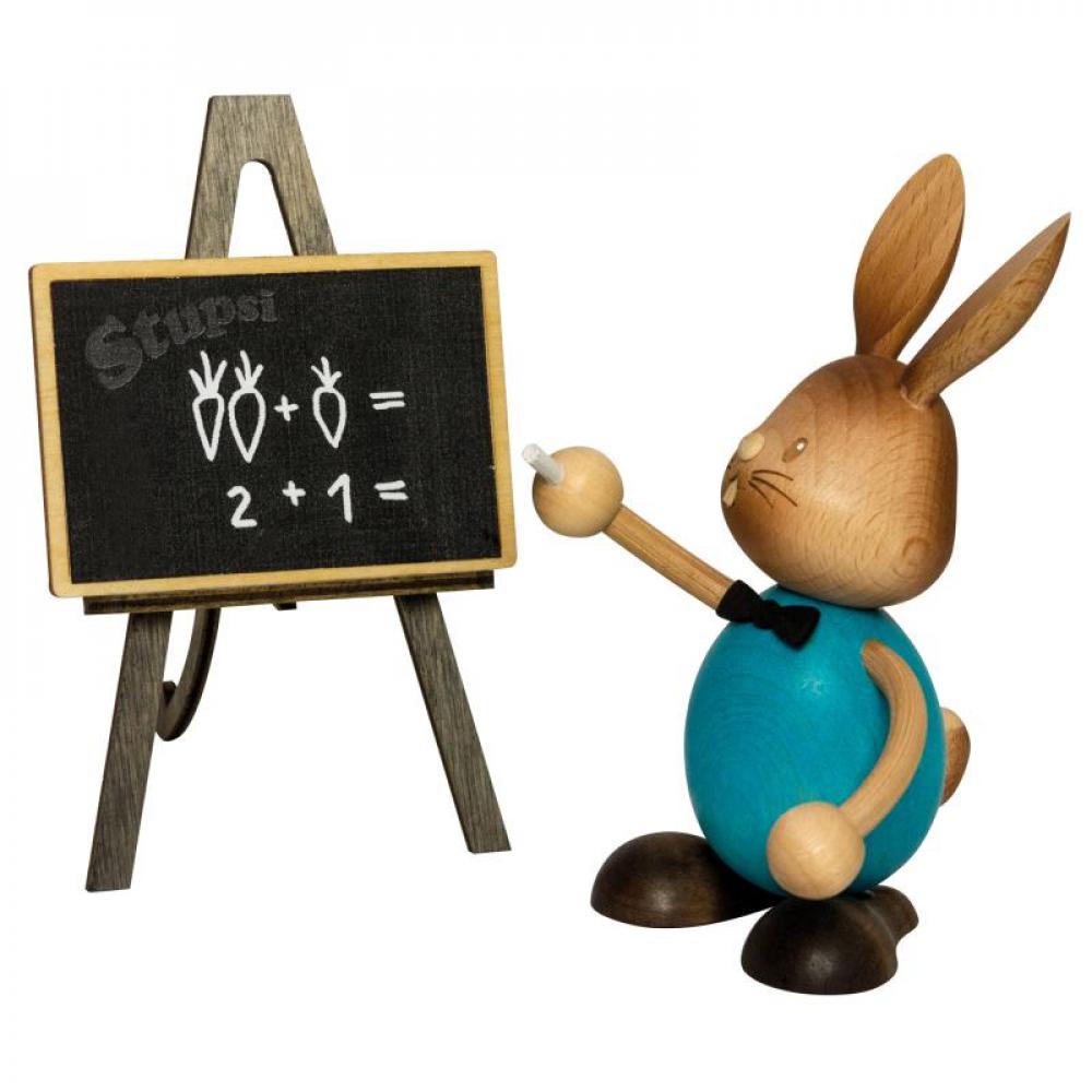 Wooden rabbit Stupsi with a blue shirt stands in front of a school blackboard and writes a math problem on it with chalk.