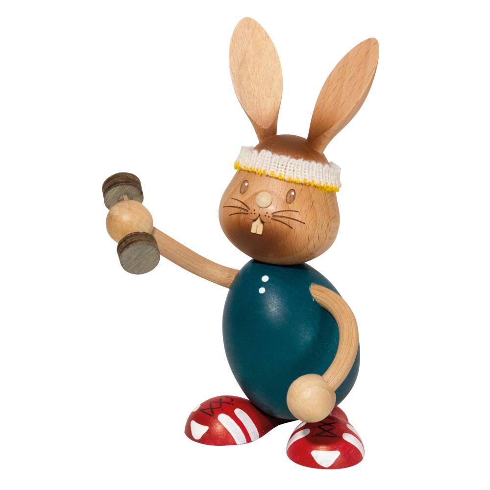 Holzhase Stupsi mit blauen Shirt schiebt eine Schubkarre vor sich her. Ein kleines Küken sitzt in der Karre und guckt heraus.